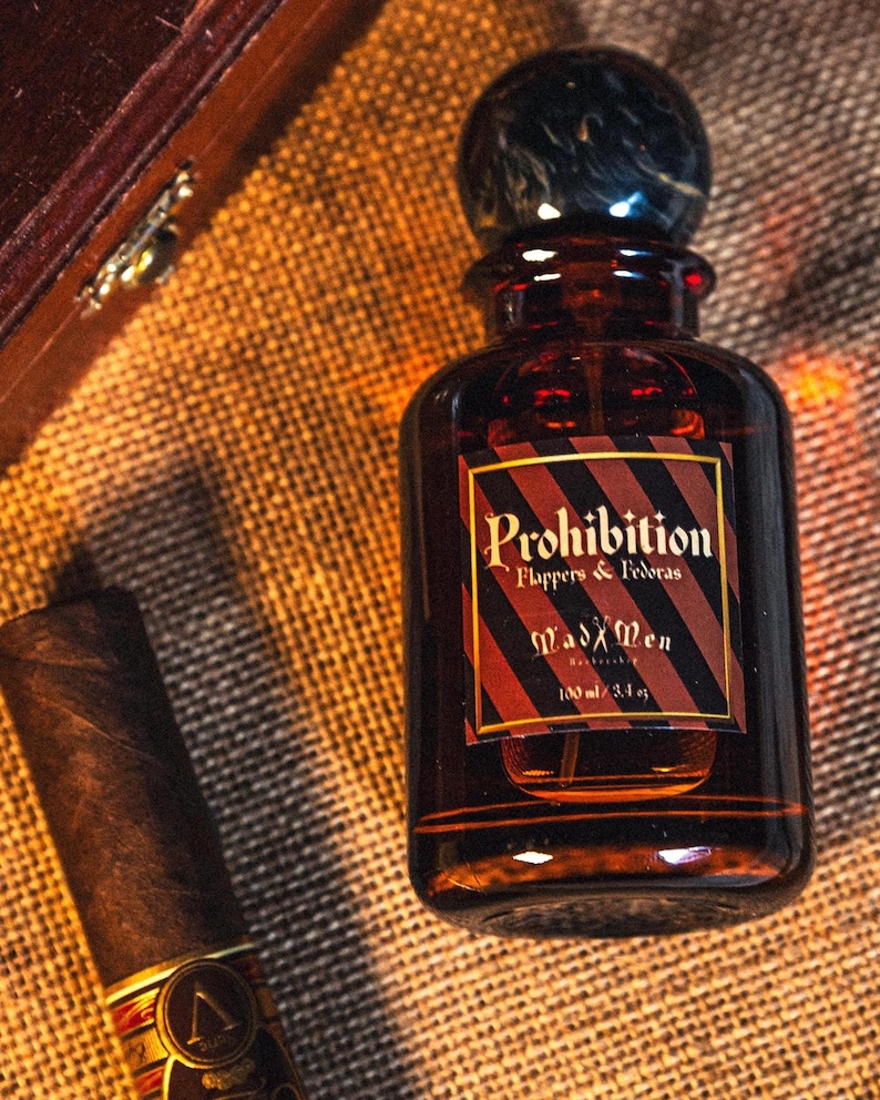 K&ouml;nnte beinhalten: Eine braune Glasflasche des Duftes "Prohibition" mit einem dunklen, marmor&auml;hnlichen Verschluss und einem Etikett mit der Aufschrift "Mad Men". Die Flasche ist 100 ml. Eine Zigarre liegt neben der Flasche auf einer strukturierten Oberfl&auml;che.