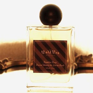 May include: A clear, square perfume bottle with a dark wooden cap. The label reads "Mad Men" and "Humber Haze, Charm Tobacco & Citrus Wood." The bottle contains 3.4 fl oz of fragrance.