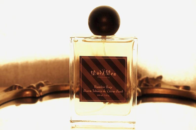May include: A clear, square perfume bottle with a dark wooden cap. The label reads "Mad Men" and "Humber Haze, Charm Tobacco & Citrus Wood." The bottle contains 3.4 fl oz of fragrance.