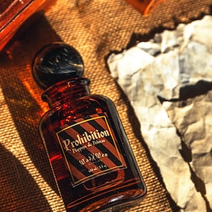K&ouml;nnte beinhalten: Eine braune Glasflasche mit einem schwarzen und braunen Verschluss. Das Etikett tr&auml;gt die Aufschrift "Prohibition Flappers & Fedoras Mad Men" mit einem diagonalen Streifendesign. Die Flasche enth&auml;lt 100 ml Fl&uuml;ssigkeit.