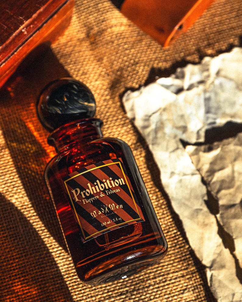 K&ouml;nnte beinhalten: Eine braune Glasflasche mit einem schwarzen und braunen Verschluss. Das Etikett tr&auml;gt die Aufschrift "Prohibition Flappers & Fedoras Mad Men" mit einem diagonalen Streifendesign. Die Flasche enth&auml;lt 100 ml Fl&uuml;ssigkeit.