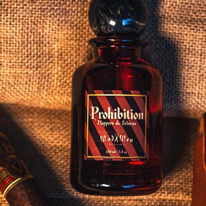 K&ouml;nnte beinhalten: Eine dunkelrote Glasflasche mit einem schwarzen, marmor&auml;hnlichen Verschluss. Das Etikett tr&auml;gt die Aufschrift "Prohibition Flappers & Fedoras" und "MadXMen" mit einem Volumen von 100 ml. Die Flasche steht vor einem strukturierten, braunen Hintergrund.