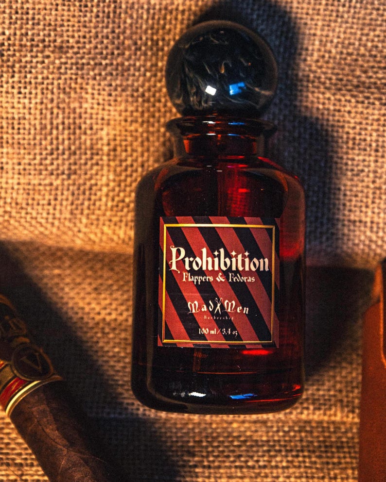 K&ouml;nnte beinhalten: Eine dunkelrote Glasflasche mit einem schwarzen, marmor&auml;hnlichen Verschluss. Das Etikett tr&auml;gt die Aufschrift "Prohibition Flappers & Fedoras" und "MadXMen" mit einem Volumen von 100 ml. Die Flasche steht vor einem strukturierten, braunen Hintergrund.