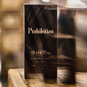 K&ouml;nnte beinhalten: Eine schwarze, rechteckige Schachtel mit "Prohibition" Eau de Parfum, diagonalen Streifen und goldfarbenen Buchstaben. Die Schachtel tr&auml;gt auch den Markennamen "Mad Men Barbershop" und 100 ml.