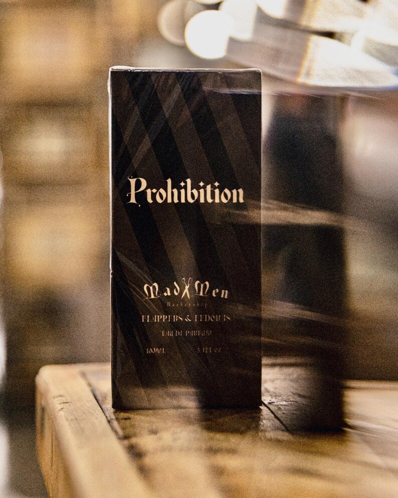 K&ouml;nnte beinhalten: Eine schwarze, rechteckige Schachtel mit "Prohibition" Eau de Parfum, diagonalen Streifen und goldfarbenen Buchstaben. Die Schachtel tr&auml;gt auch den Markennamen "Mad Men Barbershop" und 100 ml.