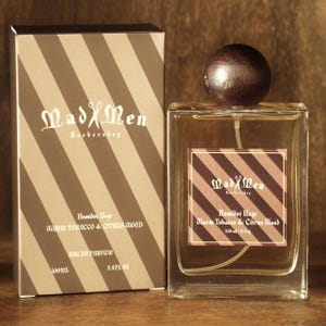 May include: A rectangular glass perfume bottle with a dark wooden cap and a matching box. The box and label feature a brown and tan striped design with the text "Mad Men Barbershop". The bottle is labeled "Humidor Haze Warm Tobacco & Citrus Wood".
