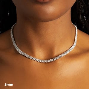 K&ouml;nnte beinhalten: Eine silberfarbene Halskette mit einer Breite von 5 mm, die ein eng verbundenes Kettendesign aufweist. Die Halskette ist mit kleinen, funkelnden Verzierungen versehen, die einen schimmernden Effekt erzeugen. Der Schmuck wird um den Hals einer Person pr&auml;sentiert.