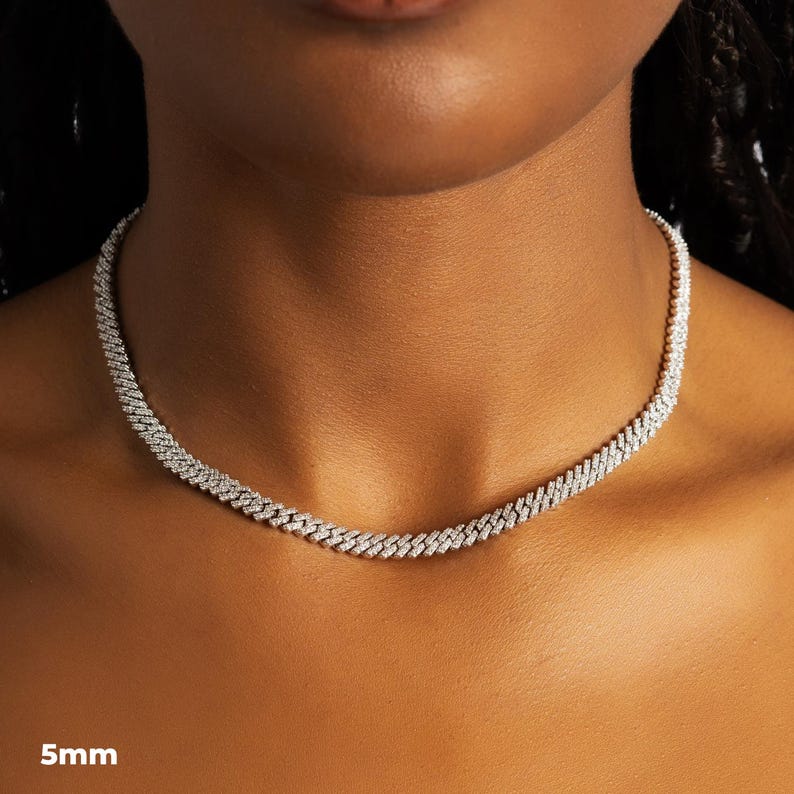 K&ouml;nnte beinhalten: Eine silberfarbene Halskette mit einer Breite von 5 mm, die ein eng verbundenes Kettendesign aufweist. Die Halskette ist mit kleinen, funkelnden Verzierungen versehen, die einen schimmernden Effekt erzeugen. Der Schmuck wird um den Hals einer Person pr&auml;sentiert.