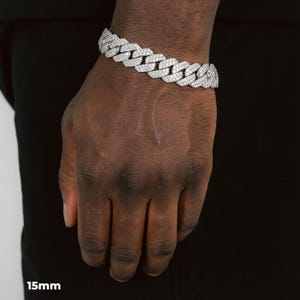 K&ouml;nnte beinhalten: Ein silberfarbenes Armband mit einem 15 mm breiten Panzerketten-Design. Das Armband ist mit zahlreichen kleinen, funkelnden Steinen besetzt, die einen schillernden Effekt erzeugen. Das Armband wird am Handgelenk einer Person vor schwarzem Hintergrund getragen.