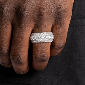 May include: A close-up of a silver ring with multiple rows of small, sparkling, clear stones. The ring is on a person's finger, with the hand and a black background visible.