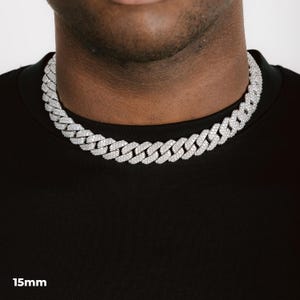 K&ouml;nnte beinhalten: Eine silberfarbene, 15 mm breite Cuban-Link-Kette. Die Halskette ist mit kleinen, funkelnden Steinen besetzt. Die Kette liegt um den Hals einer Person, vor einem schwarzen T-Shirt. Der Text "15mm" steht unten.
