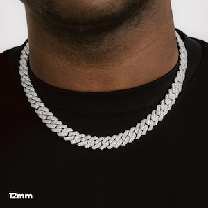 K&ouml;nnte beinhalten: Eine silberfarbene 12 mm Cuban Link-Kette. Die Halskette besteht aus kleinen, rechteckigen Gliedern, die jeweils mit kleinen, funkelnden Steinen besetzt sind. Die Halskette wird um den Hals getragen, vor einem schwarzen Hemd.