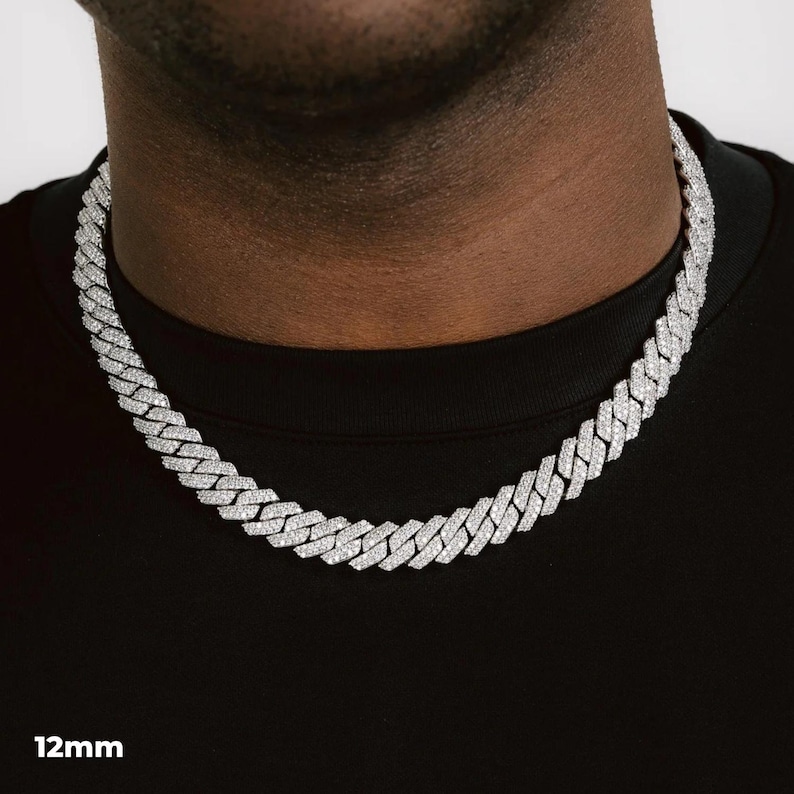 K&ouml;nnte beinhalten: Eine silberfarbene 12 mm Cuban Link-Kette. Die Halskette besteht aus kleinen, rechteckigen Gliedern, die jeweils mit kleinen, funkelnden Steinen besetzt sind. Die Halskette wird um den Hals getragen, vor einem schwarzen Hemd.