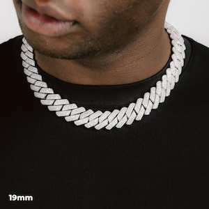 K&ouml;nnte beinhalten: Eine silberfarbene 19 mm Cuban-Link-Kette. Die Halskette besteht aus vielen kleinen, rechteckigen Gliedern, die eng miteinander verbunden sind. Die Halskette liegt um den Hals einer Person, die ein schwarzes T-Shirt tr&auml;gt.