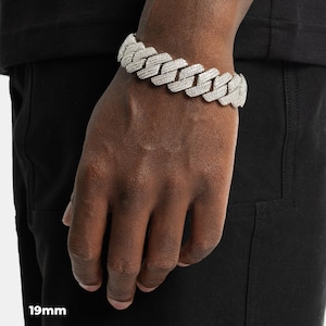 K&ouml;nnte beinhalten: Nahaufnahme eines silberfarbenen, 19 mm breiten Cuban-Link-Armbands mit einer glitzernden, strukturierten Oberfl&auml;che. Das Armband wird am Handgelenk getragen und zeigt sein kompliziertes Design und seine reflektierenden Eigenschaften. Die Details des Schmucks werden hervorgehoben.