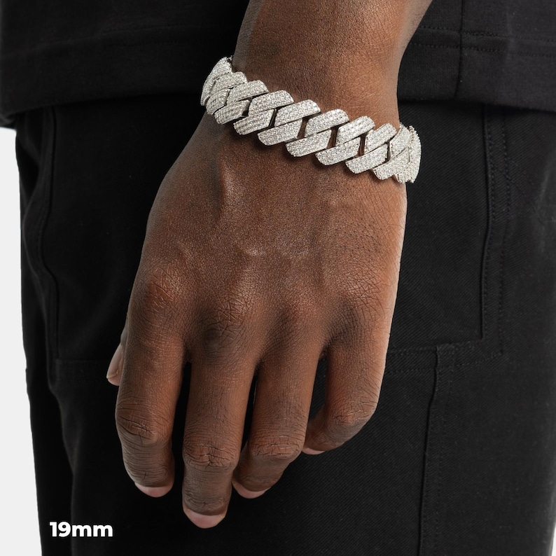 K&ouml;nnte beinhalten: Nahaufnahme eines silberfarbenen, 19 mm breiten Cuban-Link-Armbands mit einer glitzernden, strukturierten Oberfl&auml;che. Das Armband wird am Handgelenk getragen und zeigt sein kompliziertes Design und seine reflektierenden Eigenschaften. Die Details des Schmucks werden hervorgehoben.
