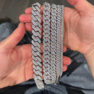 K&ouml;nnte beinhalten: Mehrere silberfarbene, mit Diamanten besetzte Kettenarmb&auml;nder in verschiedenen Breiten. Die Armb&auml;nder werden in den H&auml;nden einer Person gehalten, wodurch die komplizierten Details des Schmucks hervorgehoben werden. Die Ketten haben eine polierte, reflektierende Oberfl&auml;che.