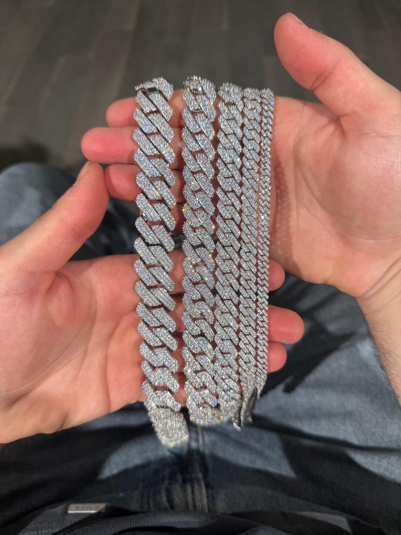 K&ouml;nnte beinhalten: Mehrere silberfarbene, mit Diamanten besetzte Kettenarmb&auml;nder in verschiedenen Breiten. Die Armb&auml;nder werden in den H&auml;nden einer Person gehalten, wodurch die komplizierten Details des Schmucks hervorgehoben werden. Die Ketten haben eine polierte, reflektierende Oberfl&auml;che.