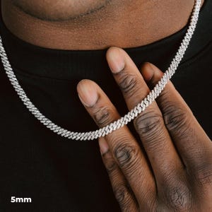 K&ouml;nnte beinhalten: Eine silberfarbene Cuban-Link-Kette mit einer Breite von 5 mm. Die Halskette besteht aus kleinen, eng aneinander liegenden, funkelnden Steinen. Die Halskette liegt um den Hals einer Person, und die Hand der Person h&auml;lt die Halskette.