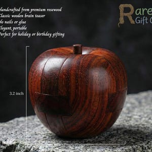 Könnte beinhalten: Ein handgefertigtes Holz-Denkspiel aus hochwertigem Rosenholz. Das Puzzle in Apfelform hat einen Durchmesser von 8,1 cm. Die Holzmaserung ist sichtbar, und das Puzzle ist ohne Nägel oder Kleber gefertigt. Das Logo von Rare Gift Co befindet sich oben rechts.
