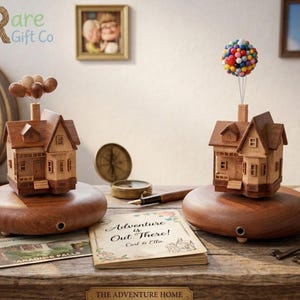 May include: Two wooden house figurines, one with brown balloons and the other with a colorful balloon cluster, sit on wooden bases. The figurines are on a wooden table with a book that reads "Adventure is Out There!" and a postcard.