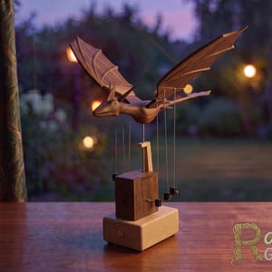 May include: A wooden automaton featuring a dark brown dragon with outstretched wings suspended above a rectangular base. The dragon is connected by thin strings to a mechanical box with a crank handle. The text Rare Gift Co is visible.