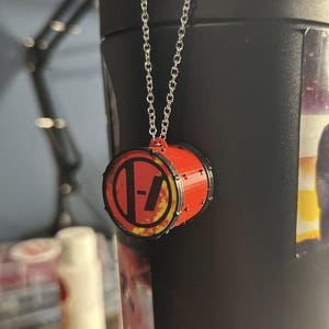 May include: A black water bottle with a silver chain and a red and yellow drum charm. The charm features a logo with a red circle, a black dash, and a yellow flame design. The bottle has a black handle and a sticker with a partial image.