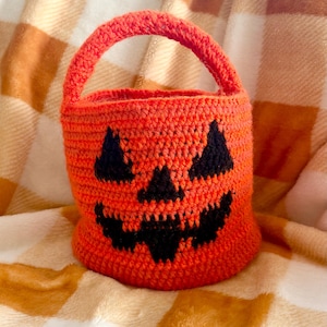 May include: An orange crocheted pumpkin-shaped basket with a black jack-o'-lantern face. The basket has a rounded base and a handle. The background is a plaid blanket in shades of orange and white.