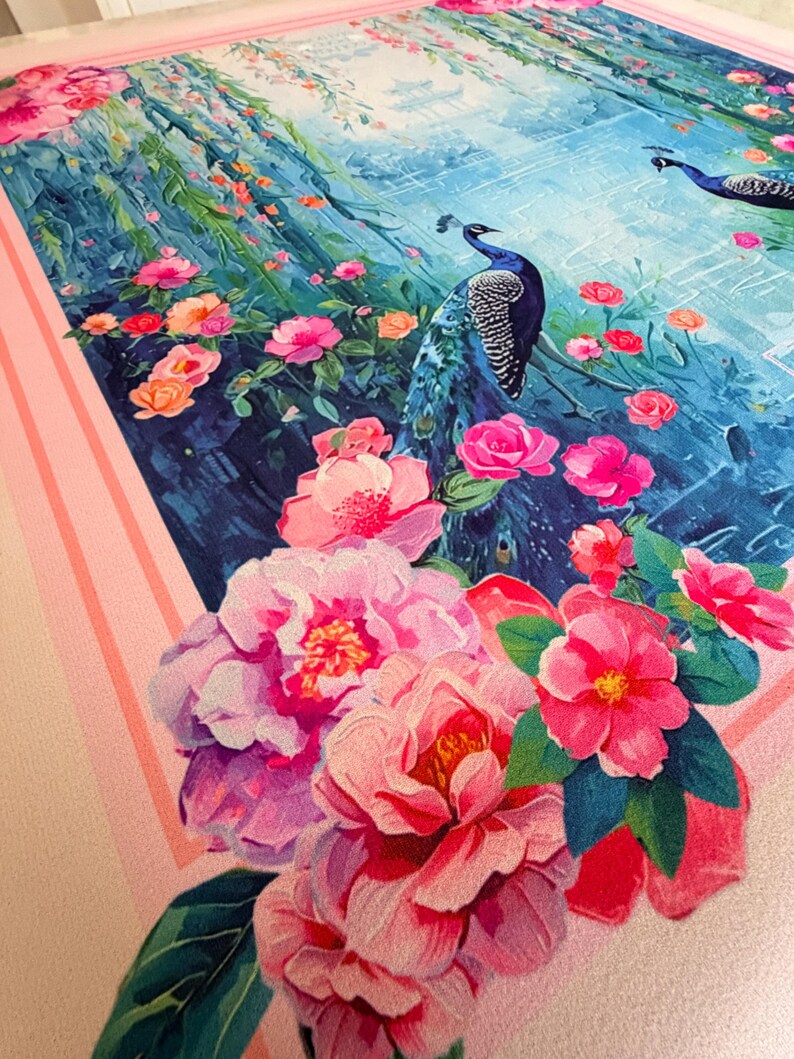 May include: A decorative textile featuring a vibrant floral and peacock design. The artwork showcases pink and red flowers, lush greenery, and two blue peacocks against a backdrop of a blue landscape. The border is a light pink with stripes.