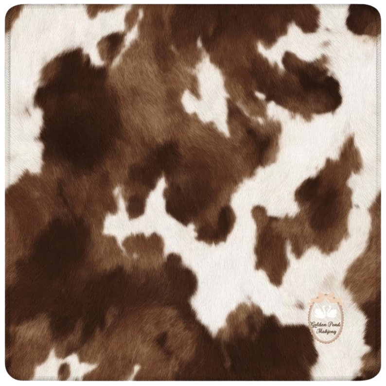 May include: A square, cowhide-patterned textile featuring a mix of brown and white patches. The design mimics the natural look of animal hide, with irregular shapes and textures. The edges are finished with a subtle, stitched border.