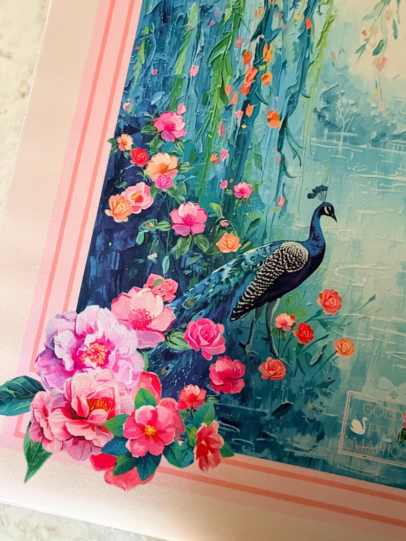 May include: A decorative scarf featuring a peacock standing amidst pink and orange flowers and lush greenery. The background depicts a blue waterfall and a serene landscape. The scarf is bordered by a pink frame with vertical stripes.