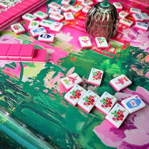 Peut inclure: Un jeu de mah-jong compos&eacute; de tuiles blanches orn&eacute;es de motifs floraux, d'oiseaux et de caract&egrave;res color&eacute;s, dispos&eacute;es sur un tapis au motif floral vert et rose vif. Une petite lampe vert fonc&eacute; est pos&eacute;e parmi les tuiles &eacute;parpill&eacute;es.