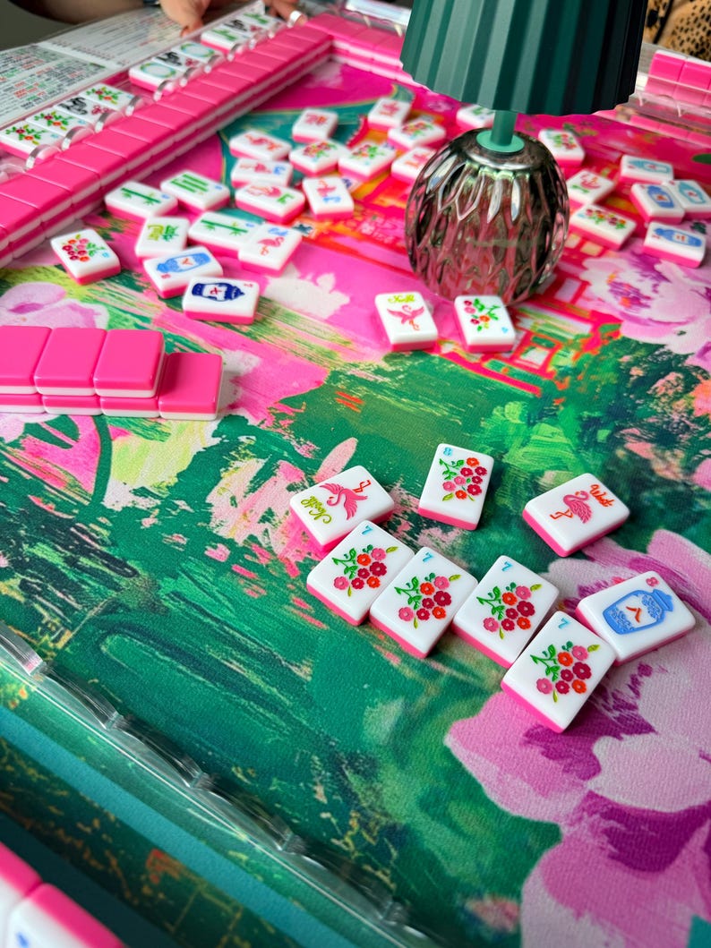 Peut inclure: Un jeu de mah-jong compos&eacute; de tuiles blanches orn&eacute;es de motifs floraux, d'oiseaux et de caract&egrave;res color&eacute;s, dispos&eacute;es sur un tapis au motif floral vert et rose vif. Une petite lampe vert fonc&eacute; est pos&eacute;e parmi les tuiles &eacute;parpill&eacute;es.