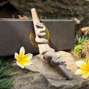 May include: A carved wooden wand featuring a detailed snake coiled around the shaft. The snake has visible scales and a defined head. The wand is positioned in front of a rectangular black box with a gold circular emblem on the front surface.