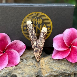 May include: A V-shaped wooden hair accessory featuring intricate pyrography patterns in dark brown on a light wood surface, positioned in front of a black box with a gold circular logo and flanked by two vibrant pink plumeria flowers.