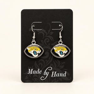 May include: A pair of silver-toned drop earrings featuring a jaguar head mascot design in yellow, black, and teal enamel. The earrings are attached to a black display card with the text Made by Hand printed in white at the bottom of the card.