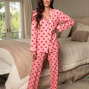 May include: Pink pajama set with a red heart pattern. The long-sleeved top has a button-down front with white trim, and the pants are straight-legged. The pajamas are made of a soft material, perfect for lounging.