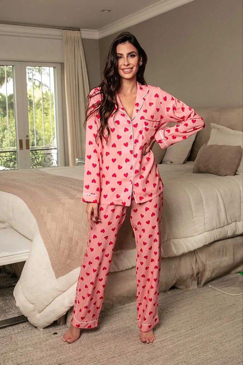 May include: Pink pajama set with a red heart pattern. The long-sleeved top has a button-down front with white trim, and the pants are straight-legged. The pajamas are made of a soft material, perfect for lounging.