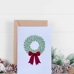 May include: A Christmas greeting card featuring a green wreath with a red bow. The card is on a white background with a brown envelope behind it. Pine cones and greenery are in the foreground.