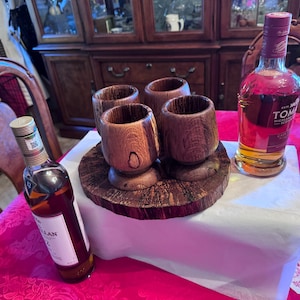May include: A display of four wooden shot glasses on a wooden base, with two bottles of liquor. One bottle is labeled "The Macallan 12", and the other is labeled "TOMINTOUL". The glasses and base are made of a dark wood.