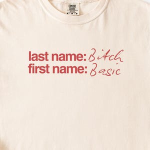 May include: Cream-colored t-shirt with a crew neck. The shirt features the text "last name: Bitch" and "first name: Basic" in a red, handwritten-style font. The shirt has a Comfort Colors tag.