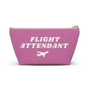 Könnte beinhalten: Rosa Kosmetiktasche mit Reißverschluss mit dem Text "FLIGHT ATTENDANT" in Weiß und einem weißen Flugzeug.