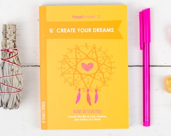 Puede incluir: Un diario amarillo y naranja con el texto "CREATE YOUR DREAMS" y "MENT ACTION PAD" en la portada. El diario está rodeado de un palo de salvia, un bolígrafo rosa, un cristal rosa y dos cristales amarillos sobre una superficie de madera blanca.