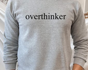 May include: Sport Grey sweatshirt with the word "overthinker" printed in black. The long-sleeved crew neck sweatshirt is a light gray color.