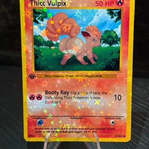 May include: A holographic trading card featuring a cartoon Vulpix character with the text "Thicc Vulpix." The card has a gold border and includes game information such as HP, attack details, and a description. The card is displayed on a clear stand.