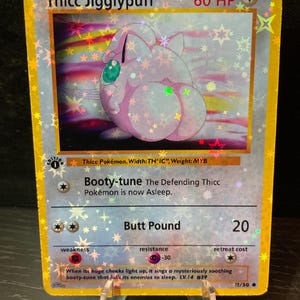 Puede incluir: Una tarjeta coleccionable holográfica con "Thicc Jigglypuff". La tarjeta tiene un borde dorado e incluye texto como "Booty-tune" y "Butt Pound". La ilustración representa un personaje rosa con un trasero grande.