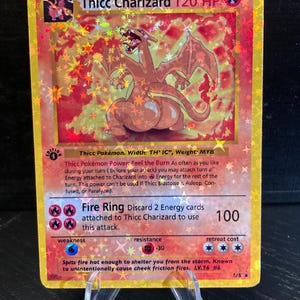 May include: A holographic Thicc Charizard Pokémon card. The card features a red and orange Charizard illustration with the text "Thicc Charizard 120 HP". The card is displayed on a clear plastic stand.