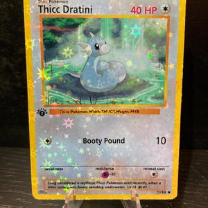 May include: A holographic trading card featuring a blue cartoon dragon named "Thicc Dratini." The card has a yellow border and includes text such as "Booty Pound" and "40 HP." The card is displayed on a clear plastic stand.