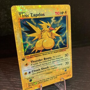 Puede incluir: Una tarjeta comercial hologr&aacute;fica dorada con un p&aacute;jaro de dibujos animados con el texto "Thicc Zapdos". La tarjeta tiene un esquema de color amarillo y azul con texto que dice "Thunder Booty" y "Cheek Shock". La tarjeta se muestra en un soporte de pl&aacute;stico transparente.