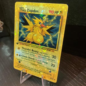 Puede incluir: Una tarjeta coleccionable hologr&aacute;fica con "Thicc Zapdos" con una ilustraci&oacute;n amarilla y azul. La tarjeta muestra texto que incluye "Thunder Booty" y "Cheek Shock" con 110 HP. La tarjeta est&aacute; apoyada en un soporte de pl&aacute;stico transparente.